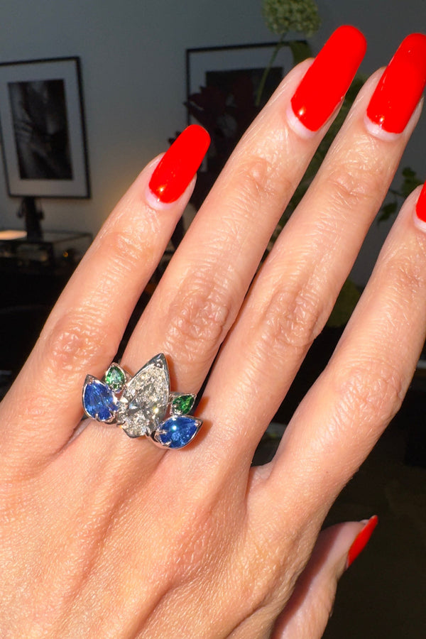 Close-up of a hand wearing a diamond ring with sapphire and emerald stones, against a blurred indoor background.